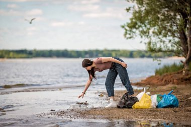 Kız sahilde çöp topluyor. Kız çöpleri kumsaldan temizliyor.