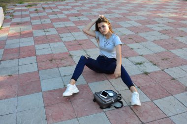 Genç, neşeli, genç bir bayan. Kırsalda oturuyor. Şık yaz kıyafetleri giyiyor. Çanta. Sıcak akşam renkleri. Güneş