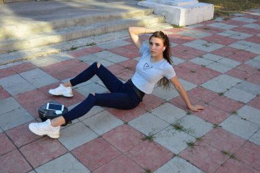 Genç, neşeli, genç bir bayan. Kırsalda oturuyor. Şık yaz kıyafetleri giyiyor. Çanta. Sıcak akşam renkleri. Güneş