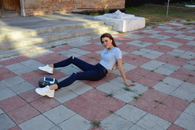 Genç, neşeli, genç bir bayan. Kırsalda oturuyor. Şık yaz kıyafetleri giyiyor. Çanta. Sıcak akşam renkleri. Güneş