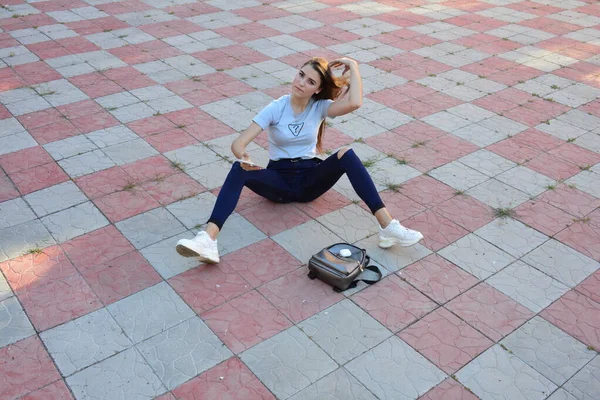 Genç, neşeli, genç bir bayan. Kırsalda oturuyor. Şık yaz kıyafetleri giyiyor. Çanta. Sıcak akşam renkleri. Güneş