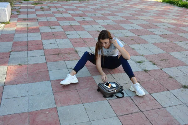 Young fresh cheerful hipster young woman sitting on the road in countryside wearing stylish summer clothes. bag . Warm evening colors. Sun