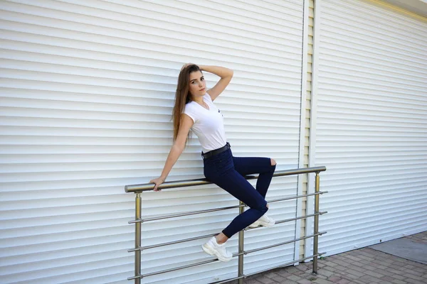 漂亮的年轻女人穿着牛仔裤 白色的T恤 站在街上 靠近铁栅栏的照片 — 图库照片