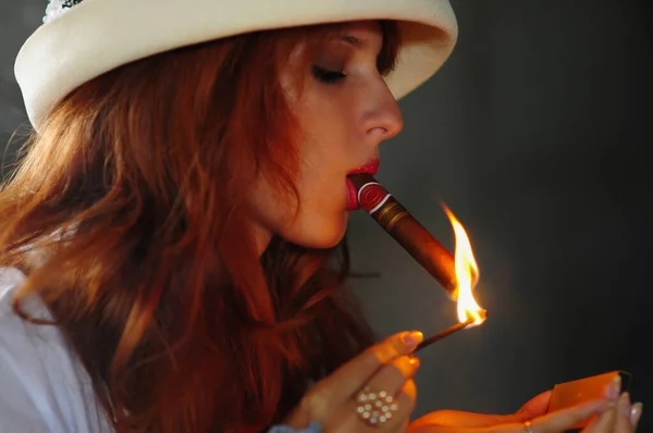 woman lit a match and lights a cigar. Elegant smoking woman. Portrait of a red-haired girl with a cigar.
