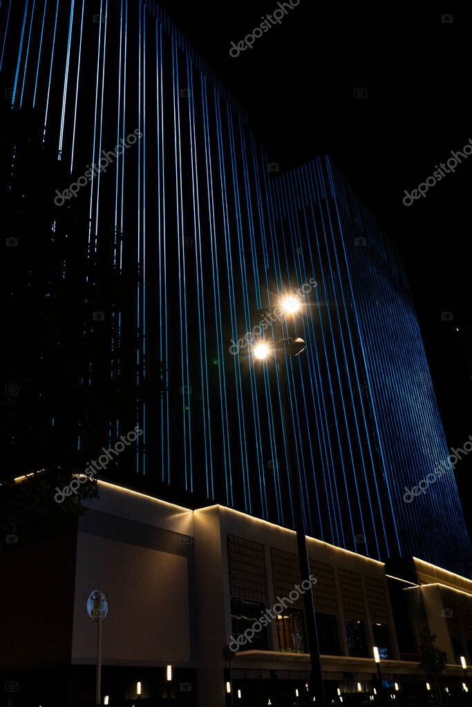 Tashkent, Uzbekistan, August 18, 2025: Sleek modern skyscraper illuminated with vertical blue and white LED lights against a dark night sky, with warm streetlights in the foreground