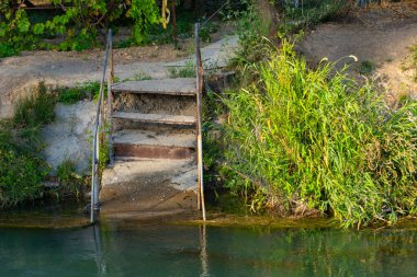 Sakin bir nehre giden eski metal ve beton merdivenler. Etrafı yaz yeşillikleriyle çevrili. Kır manzarası, dokulu adımlar ve berrak su yansıması