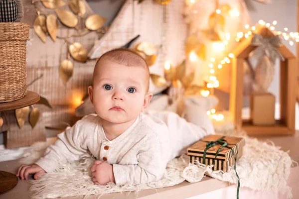 Um pequeno bebê em roupas leves fica no fundo da decoração de Natal . — Fotografia de Stock