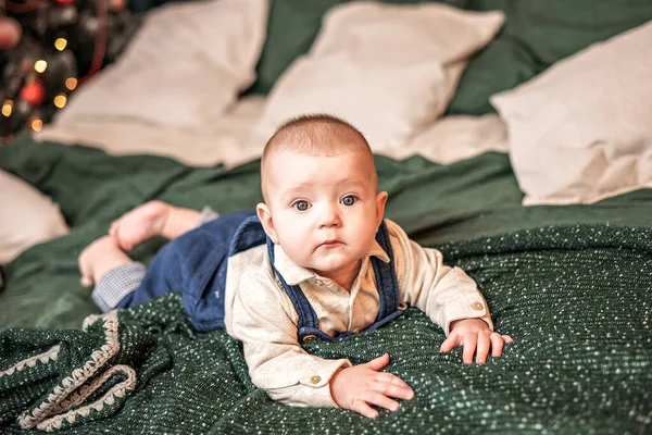 Um menino pequeno está deitado em seu estômago na cama em uma colcha verde em uma camisa clara e macacão azul . — Fotografia de Stock