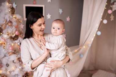 Anne, oğlunun kollarında hafif giysiler içinde Noel dekorunun arka planında..