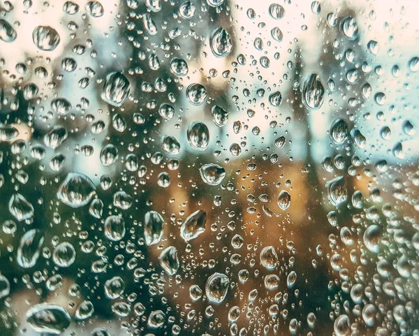 Foco seletivo. Gotas de chuva na janela de vidro depois de uma chuva de verão em um fundo com uma árvore verde, céu azul e raios de sol ao pôr do sol . — Fotografia de Stock