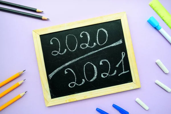 Inscriptie 2020 2021 krijt op een zwart krijtbord met een houten lijst op een paarse achtergrond en briefpapier. — Stockfoto