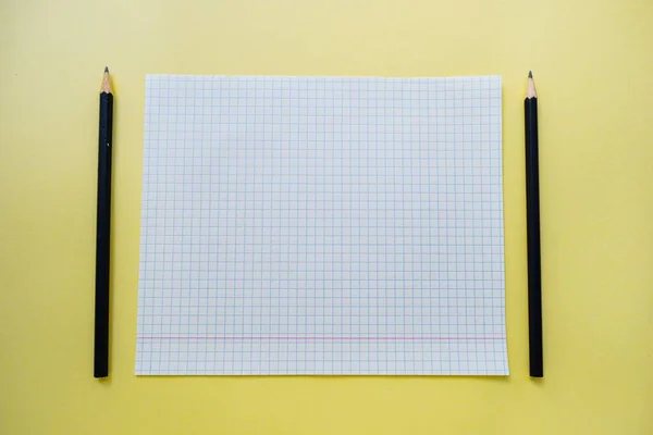 A white sheet of notebook paper in a cage on a bright yellow background with a black slate pencil. — Stock Photo, Image