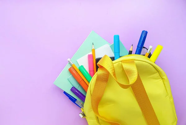 Szkolny żółty plecak z notatnikami i materiałami szkolnymi na fioletowym tle. Wracamy do szkoły.. — Zdjęcie stockowe