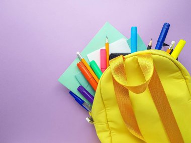 Okul sarı sırt çantası ve mor arka planda not defterleri ve okul malzemeleri. Okula geri dön..