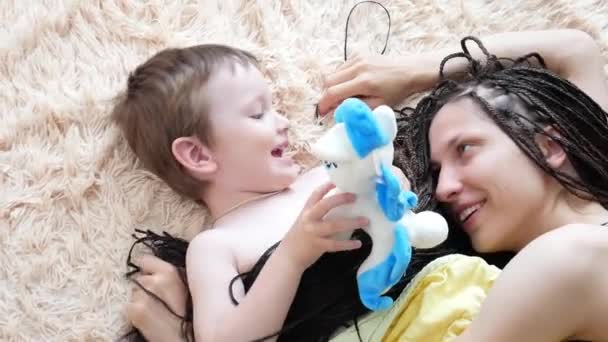 一个可爱的小男孩用他的妈妈的辫子盖住自己 和她躺在柔软的米黄色格子布上玩乐 — 图库视频影像