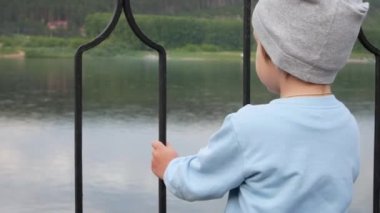 Sevimli bir çocuk ellerini çitin üzerine koyar ve nehre bakar.