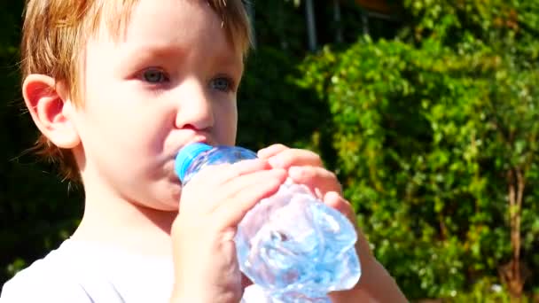 Маленький Мальчик Пьет Воду Голубой Пластиковой Бутылки Против Зеленых Зарослей — стоковое видео