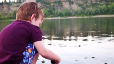 Küçük bir çocuk nehrin kıyısında otururken suya taş atıyor.