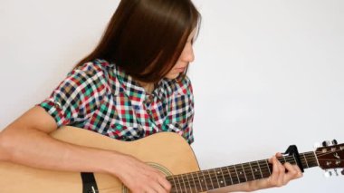 Renkli kareli gömlek giymiş güzel bir bayan beyaz arka planda akustik gitar çalıyor.