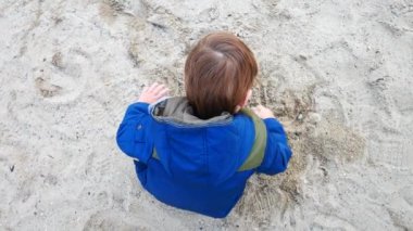 Ceket giyen küçük bir çocuk kumla oynuyor.