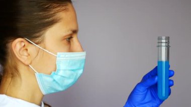 Koruyucu maskeli güzel bir laboratuvar asistanı elinde mavi sıvıyla bir test tüpü tutuyor.