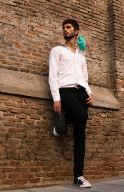 Beyaz gömlekli, eski bir tuğla duvarın önünde maske takan genç bir çocuk.