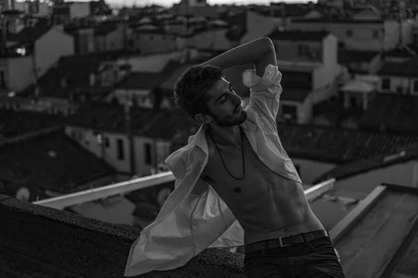 Eşcinsel çocuk beyaz bir tişörtle çatıda poz veriyor düğmeleri açık ve anın tadını çıkarıyor.