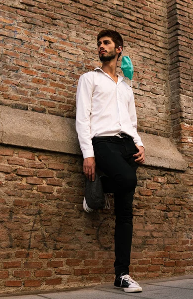 Beyaz gömlekli, eski bir tuğla duvarın önünde maske takan genç bir çocuk.