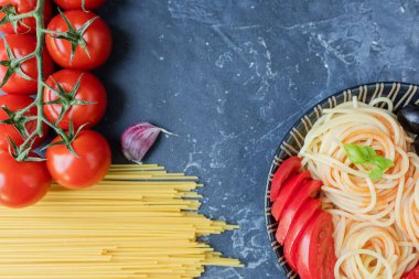 Buğday ve olgun vişneli domateslerden yapılmış spagetti bir dalda, sarımsak ve baharatlarla. Koyu grafit arkaplanda taze yeşiller. Lezzetli makarna yapmak için ürünler. Yukarıdan görünüşü.