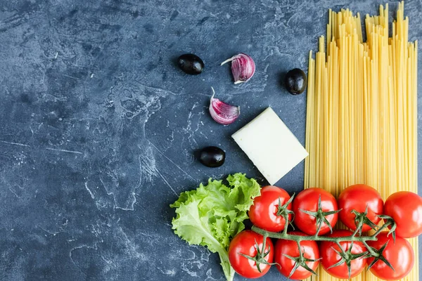Buğday ve olgun vişneli domateslerden yapılmış spagetti bir dalda, sarımsak ve baharatlarla. Koyu grafit arkaplanda taze yeşiller. Lezzetli makarna yapmak için ürünler. Yukarıdan görünüşü.