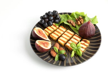 Koyu renkli, yuvarlak bir tabakta kızarmış halloumi peyniri, beyaz arka planda meyve, fındık ve salata yaprakları. Yunanistan ve Türkiye 'nin geleneksel peyniri. Boşluğu kopyala. 