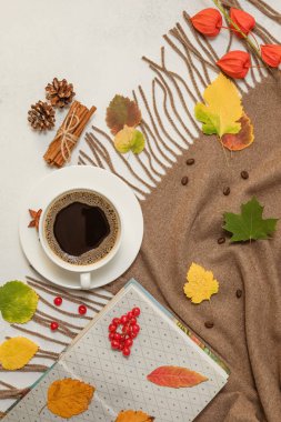 Sonbahar kompozisyonu. Bardakta kahve, kahverengi eşarp, kitap, tarçın çubukları, beyaz bir masanın üzerine düşen yapraklar. Seçici odak.