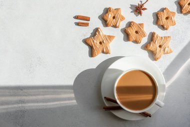 Beyaz eskimiş arka planda, yıldız şeklinde ev yapımı kurabiye ve beyaz porselen bir fincanda sütlü Noel kahvesi. Yukarıdan manzara. sert güneş ışığı