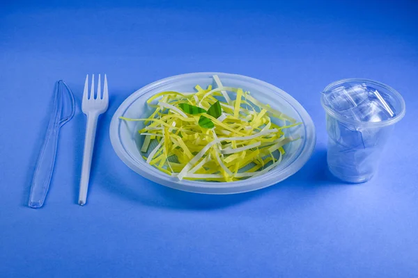 Disposable plastic tableware, plate, fork, knife, glass on a blue background. The plate contains plastic food. Composition for environmentalists. Healthy nutrition.