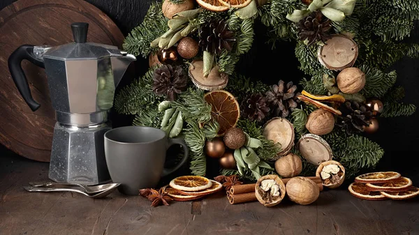 Christmas mood composition with wreath, coffee pot, cup, walnuts and dried orange slices. Front view on a dark wooden background