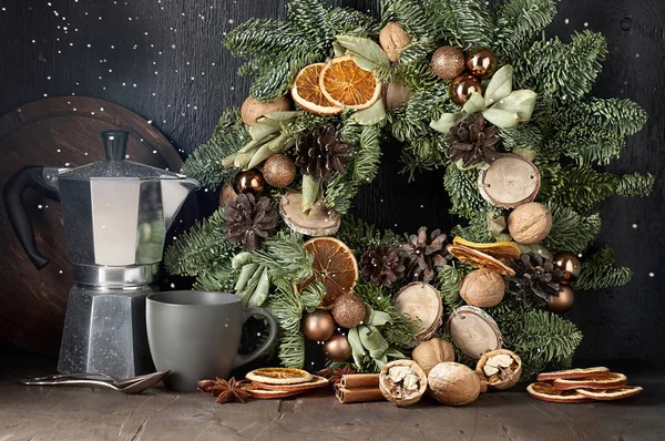 Christmas mood composition with wreath, coffee pot, cup, walnuts, dried orange slices and falling snow. Front view on a dark wooden background