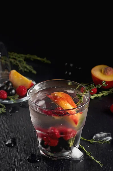 Raspberry thyme gin and tonic cocktail with blackcurrant and peaches. Refreshing summer drink with ice on black background. Close up view with copy space