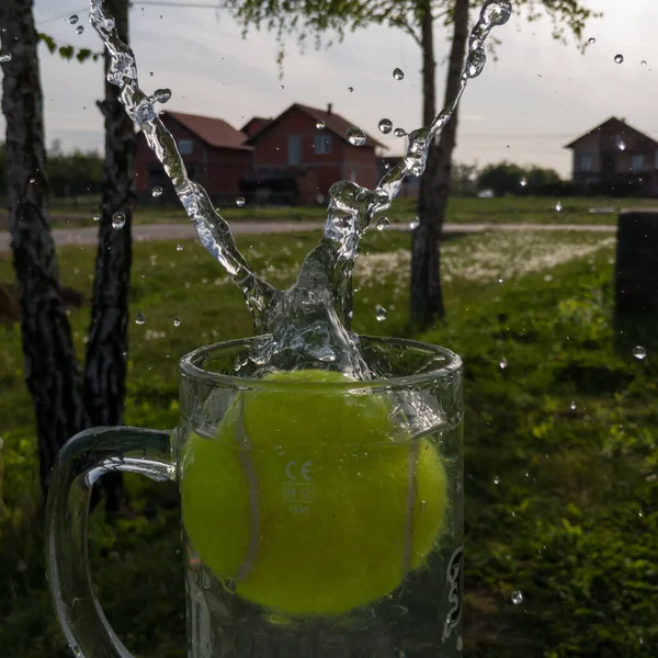 Tennis ball make water splash from glass mug against countryside during sunny day