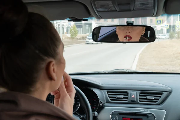 Young female driver make-up in the car looking at rearview mirror. Young woman painting bright red lipstick on her lips.