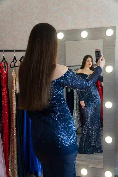 Belle jeune femme en robe bleu foncé étincelante debout devant le miroir faisant selfie. Concentre-toi sur le miroir. Robes de luxe sur les cintres au magasin de location pour bal, mariage ou séance photo. Images De Stock Libres De Droits