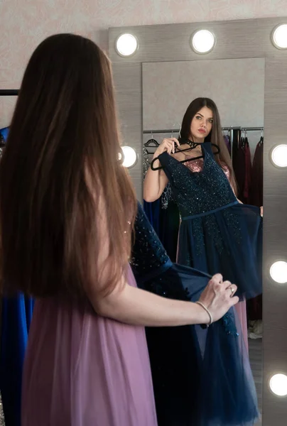 Belle jeune femme brune debout devant le miroir en choisissant une robe glamour. Concentre-toi sur le miroir. Robe de luxe dans le magasin de location pour bal, mariage ou photo. Images De Stock Libres De Droits