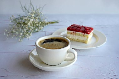 Beyaz arka planda beyaz kahve fincanı, beyaz tabakta tatlı, arka planda çiçekler.                         