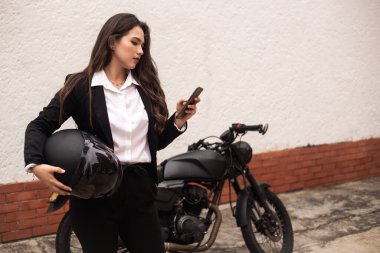 Motosikletine binmeden önce cep telefonunu kullanan bir kadın. Bir kask tutuyor.