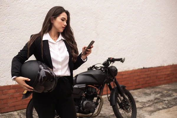 Motosikletine binmeden önce cep telefonunu kullanan bir kadın. Bir kask tutuyor.