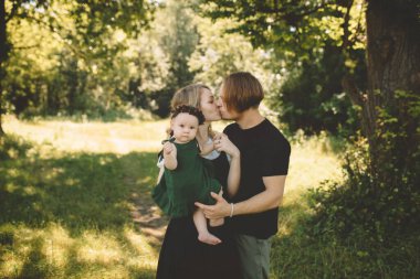 Anne ve baba küçük kızlarını kucağına alıp parkta öpüşüyorlar. Mutlu aile ve çocukluk