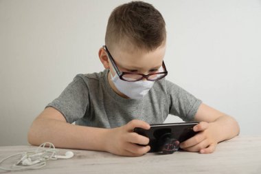 Gri tişörtlü bir çocuk telefonun başına oturur ve internete bağlanır, uzaktan öğrenme, telefon başında masa baskısı alan bir çocuk telefona bakar.. 