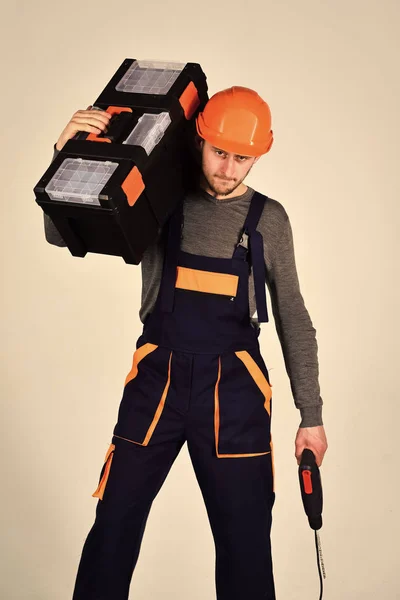 Klusjesman concept. Man in overall en helm houdt toolbox op schouder en boor in de hand. Sterke en aantrekkelijke hersteller, grijze achtergrond, geïsoleerd. Macho op geconcentreerde gezicht draagt tools voor reparatie. — Stockfoto