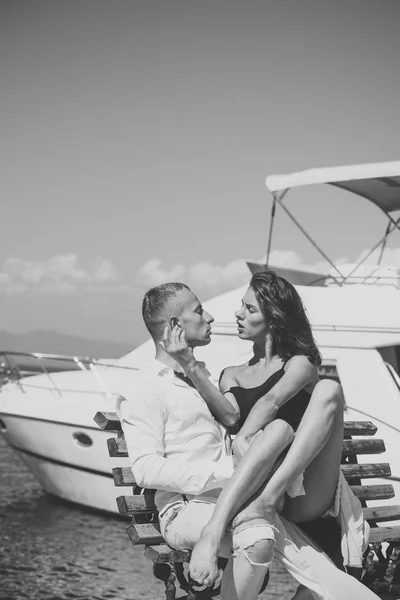Couple passionné assis sur le banc au quai près du bateau et câlins, journée d'été ensoleillée. Femme et homme en vêtements à la mode pleins de désir près d'un yacht de luxe. Concept de lune de miel. Couple amoureux en lune de miel . — Photo