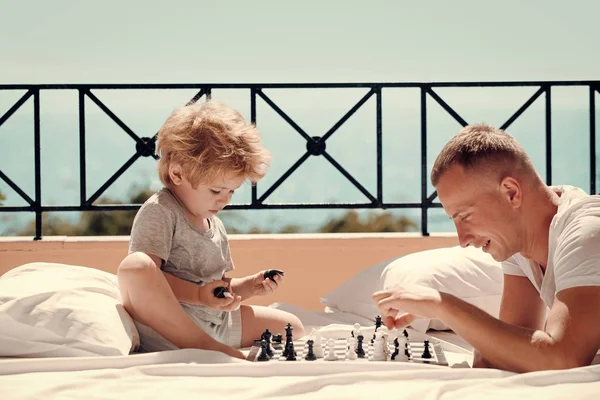 Concetto di bambino intelligente. I genitori giocano a scacchi con il bambino sulla terrazza nella giornata di sole. Papà con bambino gioco intellettuale. Padre e figlioletto giocano a scacchi su balcone, natura e mare sullo sfondo . — Foto Stock