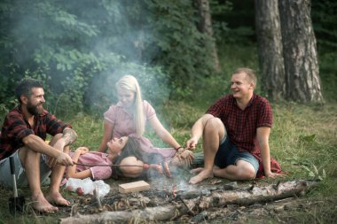 Ormanda piknik arkadaşları gülümseyerek. İki mutlu çiftler zevk yaz akşam açık havada, romantik tarih. Sakallı adam ve arkadaşı arkadaşlarını dinlenme süre ateşte Sosis kızartma.
